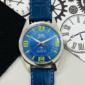 May include: A silver HMT Jawan wristwatch with a blue face and green accents. The watch has a blue leather strap and says "17 Jewels Para Shock" and "Made in India 0231".
