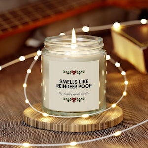 May include: A lit glass jar candle on a wooden coaster. The candle has a white label with the text "Smells Like Reindeer Poop" and "My Holiday Spirit Candle". The candle is surrounded by a string of lights.
