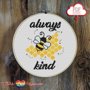Può includere: Un cerchio da ricamo bianco con un disegno a punto croce di un'ape e un favo con il testo "always be kind".