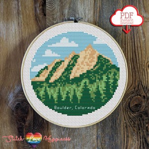 以下が含まれることがあります： 青い空と雲のある山の風景を描いたクロスステッチ刺繍枠。「Boulder, Colorado」の文字が下部に刺繍されています。 「PDF Instant Download」と書かれた赤い雲のグラフィックが右上隅にあります。