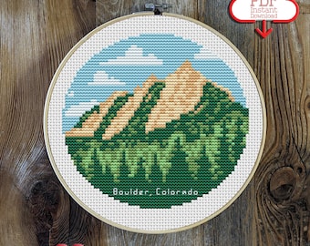 May include: A cross-stitch embroidery hoop featuring a mountain landscape with a blue sky and white clouds. The text "Boulder, Colorado" is stitched at the bottom. A red cloud graphic with "PDF Instant Download" is in the upper right.