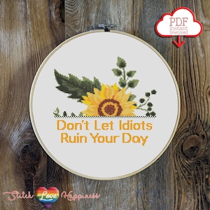 May include: A circular embroidery hoop containing a cross-stitch pattern of a yellow sunflower with green leaves and stems. Below the flower, the text reads, "Don't Let Idiots Ruin Your Day" in orange letters. A red cloud icon with "PDF Instant Download" is visible.