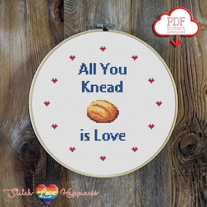 Puede incluir: Un aro de bordado blanco con un diseño de punto de cruz que dice "All You Knead is Love" con una ilustración de pan marrón. El aro está rodeado de corazones rojos.