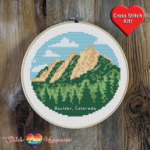 May include: A cross-stitch kit featuring a mountain landscape with a blue sky and clouds. The design includes green trees and tan mountains, with the text "Boulder, Colorado." A red heart-shaped tag reads "Cross Stitch Kit!"