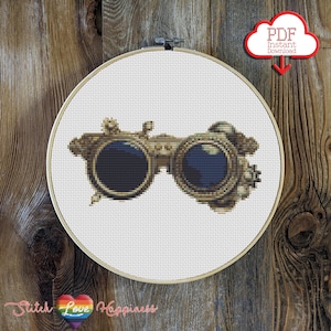 May include: A cross stitch pattern featuring a pair of vintage steampunk goggles with gold accents and blue lenses. The pattern is displayed in a round embroidery hoop.