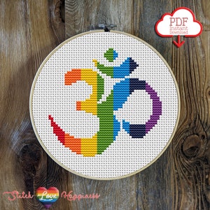 Peut inclure: Un motif de point de croix représentant un symbole Om de couleur arc-en-ciel dans un cercle de broderie blanc. Le motif est sur une toile Aida blanche. Le cercle est sur un fond en bois.