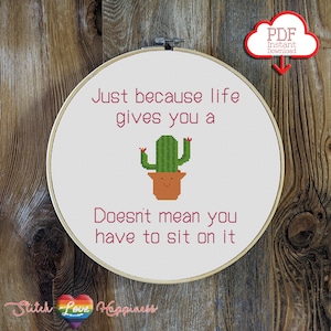 Puede incluir: Un aro de bordado blanco con un diseño de punto de cruz que presenta un cactus en una maceta y el texto "Just because life gives you a Doesn't mean you have to sit on it".
