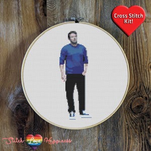 Puede incluir: Kit de punto de cruz con una imagen pixelada de una persona con camisa azul y pantalones negros, dentro de un bastidor de bordado de madera. Un corazón rojo con el texto "Cross Stitch Kit!" es visible.