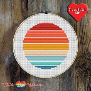 Op de afbeelding: Een kruissteekpakket met een retro zonsondergangontwerp. Het ontwerp heeft horizontale strepen in rood, oranje, geel en blauw. De kit zit in een houten hoepel, met een rood hartvormig label met de tekst "Cross Stitch Kit!"