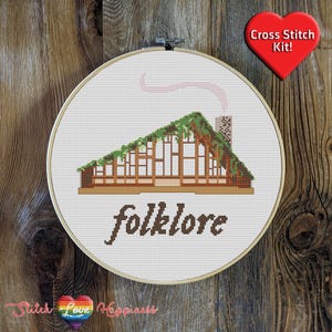 Puede incluir: Kit de punto de cruz con un fondo blanco, una casa marrón con un techo verde y la palabra "folklore" en letras marrones debajo de la casa.