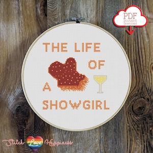 Puede incluir: Un bastidor de bordado de punto de cruz con el texto "THE LIFE OF A SHOW GIRL" en naranja. El diseño incluye un zapato de corista, un cóctel y un icono de descarga PDF. El fondo es de madera.
