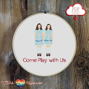 May include: Cross stitch pattern featuring two girls in blue dresses with the text "Come Play with Us" in red. The pattern is on a white background and is framed in a wooden hoop.