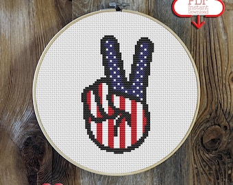 May include: Peace sign cross stitch pattern featuring an American flag design. The pattern is displayed in a white embroidery hoop against a wooden backdrop.