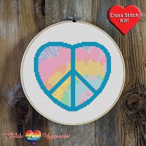May include: A cross stitch kit featuring a peace sign in a heart shape. The peace sign is filled with a tie-dye pattern in shades of pink, yellow, and blue. The kit is framed in a wooden hoop.