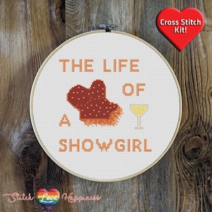 May include: A cross-stitch kit in a wooden hoop with the text "THE LIFE OF A SHOWGIRL". The design includes a heart-shaped object, a cocktail glass, and the words in orange. A red heart with "Cross Stitch Kit!" is in the upper right.