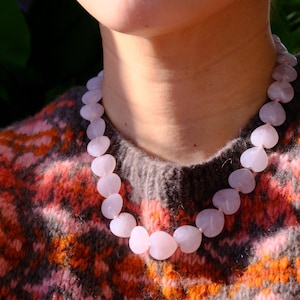 Puede incluir: Un collar de cuentas rosas claras en forma de corazón. El collar se lleva sobre un suéter con un estampado de color naranja, rosa y gris. Las cuentas son lisas y translúcidas.