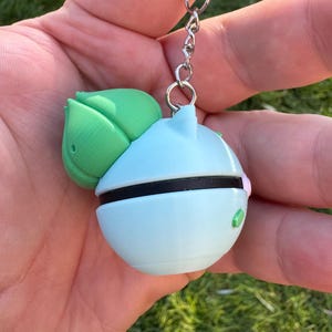 May include: A light blue and green keychain featuring a round design with a black band and a green leaf-like shape. The keychain is attached to a silver metal chain.