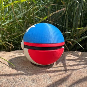May include: A 3D-printed spherical toy, featuring a blue top with white stars, a red band, and a red and white base. The toy is placed on a stone surface, with green foliage in the background. The toy is a replica of a popular animated series.