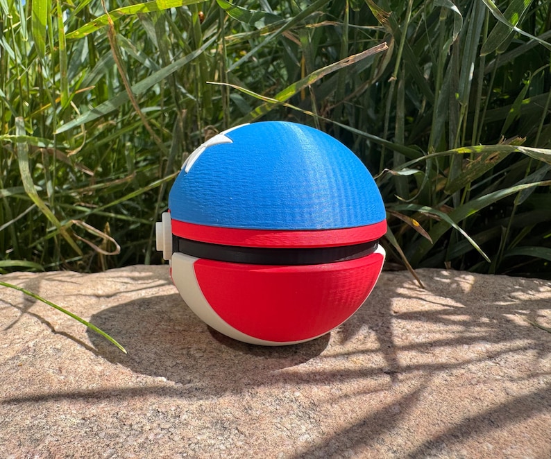 May include: A 3D-printed spherical toy, featuring a blue top with white stars, a red band, and a red and white base. The toy is placed on a stone surface, with green foliage in the background. The toy is a replica of a popular animated series.
