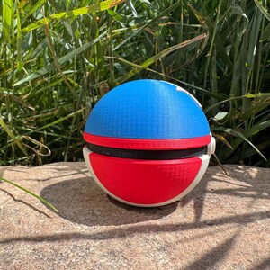 May include: A spherical object with a blue top, red bottom, and black band. The sides are white. The object is resting on a stone surface, with green foliage in the background. It appears to be a toy or decorative item.