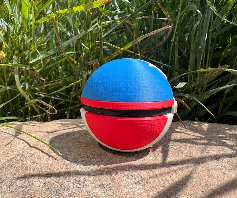 May include: A spherical object with a blue top, red bottom, and black band. The sides are white. The object is resting on a stone surface, with green foliage in the background. It appears to be a toy or decorative item.