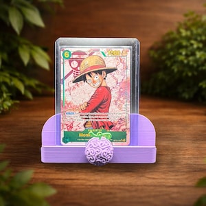 May include: A trading card featuring a character in a straw hat and red shirt, displayed in a clear protective sleeve. The card is held by a light purple stand with a decorative purple fruit. The background is a wooden surface with green foliage.