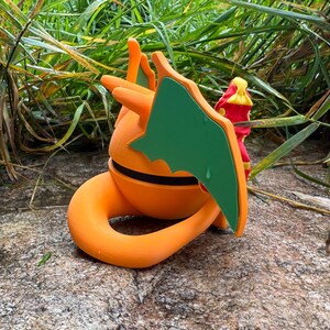 May include: A 3D-printed figure of a dragon-like creature in orange and green, with a curled tail and wings. It features a black stripe around the middle. The figure is set against a natural backdrop of green foliage and a stone surface.