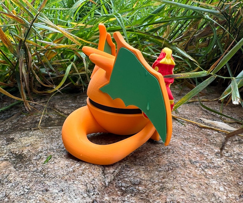 May include: A 3D-printed figure of a dragon-like creature in orange and green, with a curled tail and wings. It features a black stripe around the middle. The figure is set against a natural backdrop of green foliage and a stone surface.