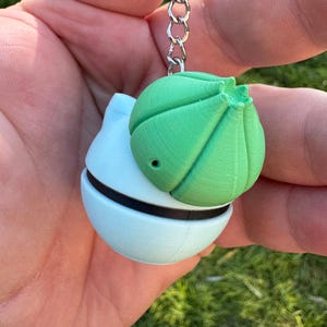 May include: A light blue and green keychain featuring a spherical design. The keychain has a green, textured top and a light blue base with a black band. The keychain is attached to a silver chain.
