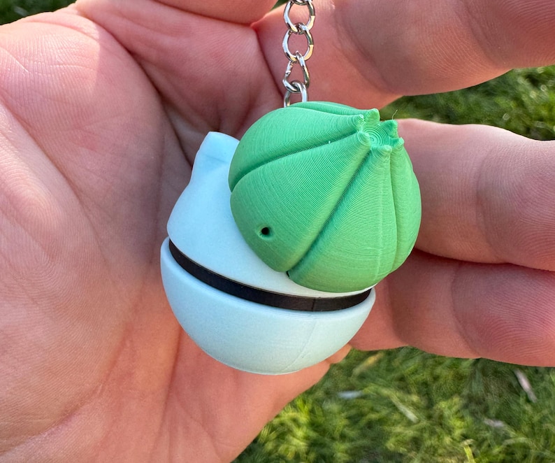 May include: A light blue and green keychain featuring a spherical design. The keychain has a green, textured top and a light blue base with a black band. The keychain is attached to a silver chain.