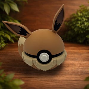 May include: A spherical figurine resembling the character Eevee, featuring a tan and cream color scheme, large pointed ears with dark brown inner details, and a central black and white circular emblem resembling a ball-shaped design.