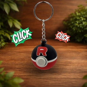 May include: A spherical keychain featuring a black and red design with a white center button and a prominent red letter R on the top half. The keychain includes a silver metal ring and chain. Two comic-style speech bubbles with the text CLICK appear.