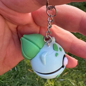 May include: A light blue Bulbasaur keychain with a green leaf and accents. The keychain is attached to a silver chain and ring. The Bulbasaur figure has a white and pink flower-like detail on its side.