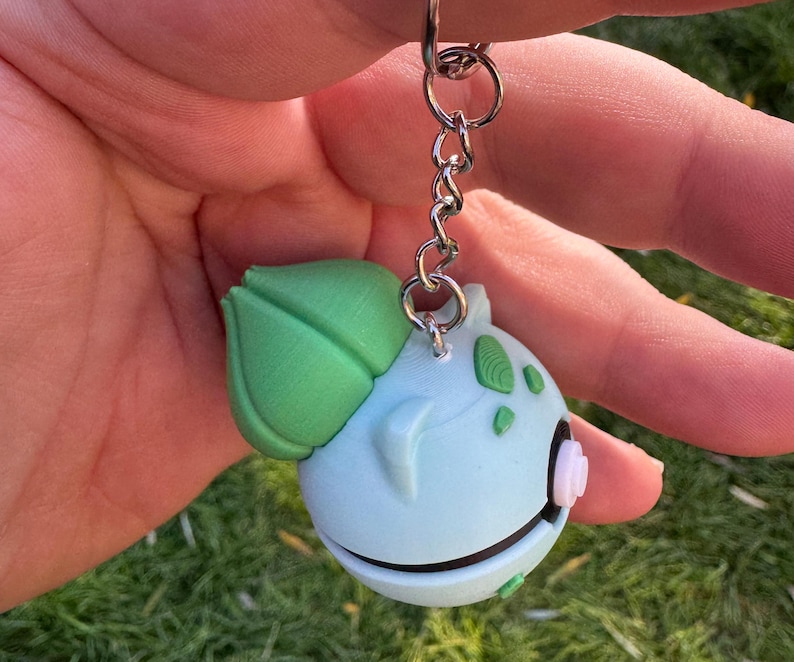 May include: A light blue Bulbasaur keychain with a green leaf and accents. The keychain is attached to a silver chain and ring. The Bulbasaur figure has a white and pink flower-like detail on its side.