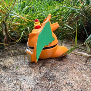 May include: An orange dragon figurine with green wings, white claws, and a red and yellow head crest. The dragon is resting on a rock, surrounded by green grass.
