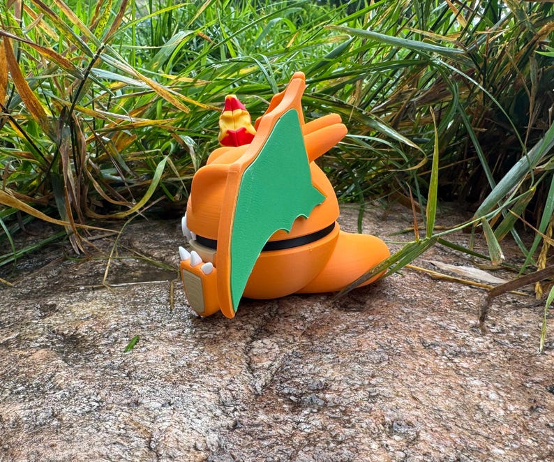 May include: An orange dragon figurine with green wings, white claws, and a red and yellow head crest. The dragon is resting on a rock, surrounded by green grass.