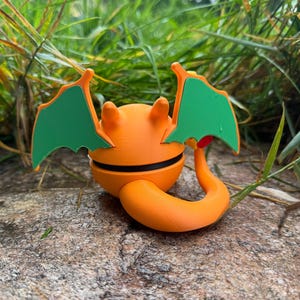 May include: An orange and black figurine with green wings and a curled tail. The figurine is resting on a rock, surrounded by green grass. The design is reminiscent of a cartoon character.