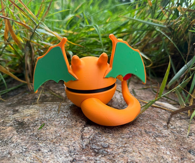 May include: An orange and black figurine with green wings and a curled tail. The figurine is resting on a rock, surrounded by green grass. The design is reminiscent of a cartoon character.