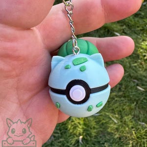 May include: A light blue and green Bulbasaur keychain. The keychain features a Bulbasaur design with a green bulb on top, light blue body, and a black and white circle in the middle. The keychain is attached to a silver chain.