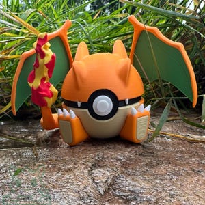 May include: A 3D-printed Charizard figure, with orange, cream, and green colours. The figure has a flame tail, wings, and a round body. The figure is sitting on a rocky surface with green foliage in the background.