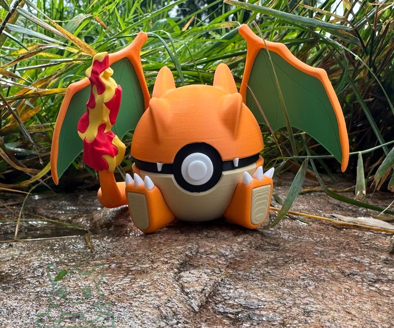 May include: A 3D-printed Charizard figure, with orange, cream, and green colours. The figure has a flame tail, wings, and a round body. The figure is sitting on a rocky surface with green foliage in the background.