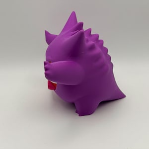 Pu&ograve; includere: Una figura tridimensionale viola e arrotondata, con orecchie a punta e una serie di punte lungo la schiena. La figura ha una bocca ampia e aperta che rivela una lingua rosso brillante ed &egrave; posizionata di profilo su uno sfondo neutro.