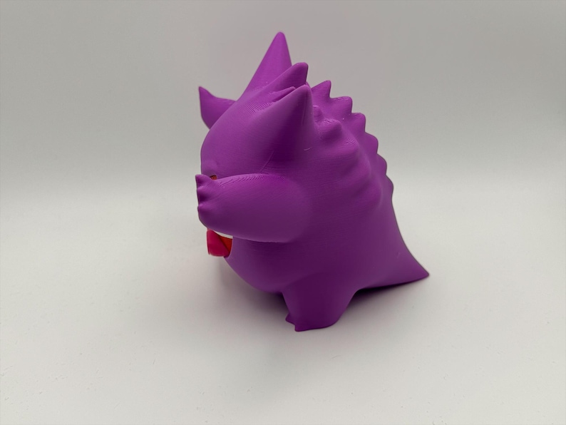 Pu&ograve; includere: Una figura tridimensionale viola e arrotondata, con orecchie a punta e una serie di punte lungo la schiena. La figura ha una bocca ampia e aperta che rivela una lingua rosso brillante ed &egrave; posizionata di profilo su uno sfondo neutro.