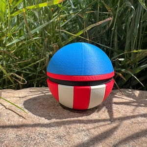 May include: A spherical, multi-coloured object with a blue top, red and black middle section, and a red and white striped bottom. The object is on a rock in front of tall green grass.