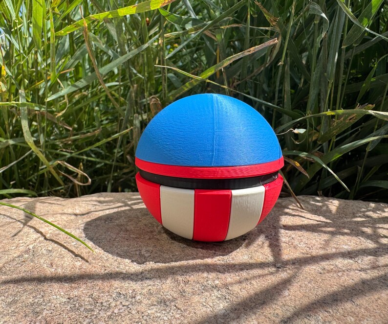 May include: A spherical, multi-coloured object with a blue top, red and black middle section, and a red and white striped bottom. The object is on a rock in front of tall green grass.