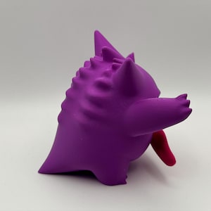 Pu&ograve; includere: Una statuetta viola di una creatura rotonda con testa appuntita, punte multiple lungo la schiena e una lunga lingua rossa sporgente. Il personaggio ha un corpo arrotondato, arti corti e una finitura opaca liscia, ripreso di profilo.