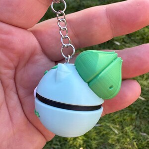 May include: A light blue and green keychain featuring a spherical design with a green leaf-like structure on top. The keychain has a black band around the middle and is attached to a silver chain.