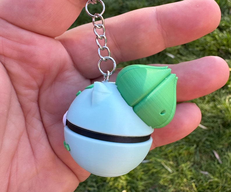 May include: A light blue and green keychain featuring a spherical design with a green leaf-like structure on top. The keychain has a black band around the middle and is attached to a silver chain.