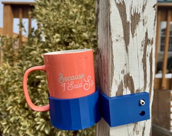 May include: A blue plastic mug holder attached to a wooden post. A coral colored mug with the text "Because I Said So" is in the holder.