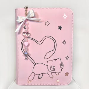 May include: Pink binder featuring a black cat outline with a heart-shaped tail. It has a charm with a white bow, beads, and a small decorative piece. Stars and other shapes are scattered across the front.